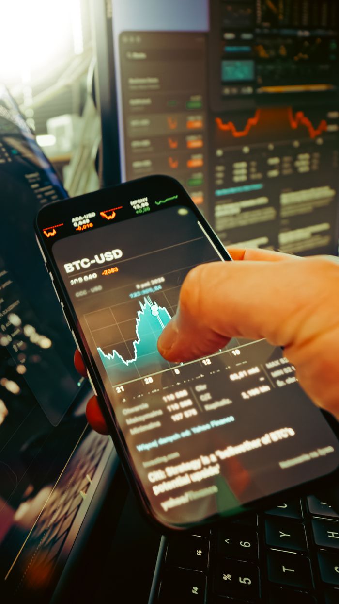 gallery-01 A hand holds a smartphone displaying BTC-USD chart amidst digital trading screens, highlighting crypto finance.